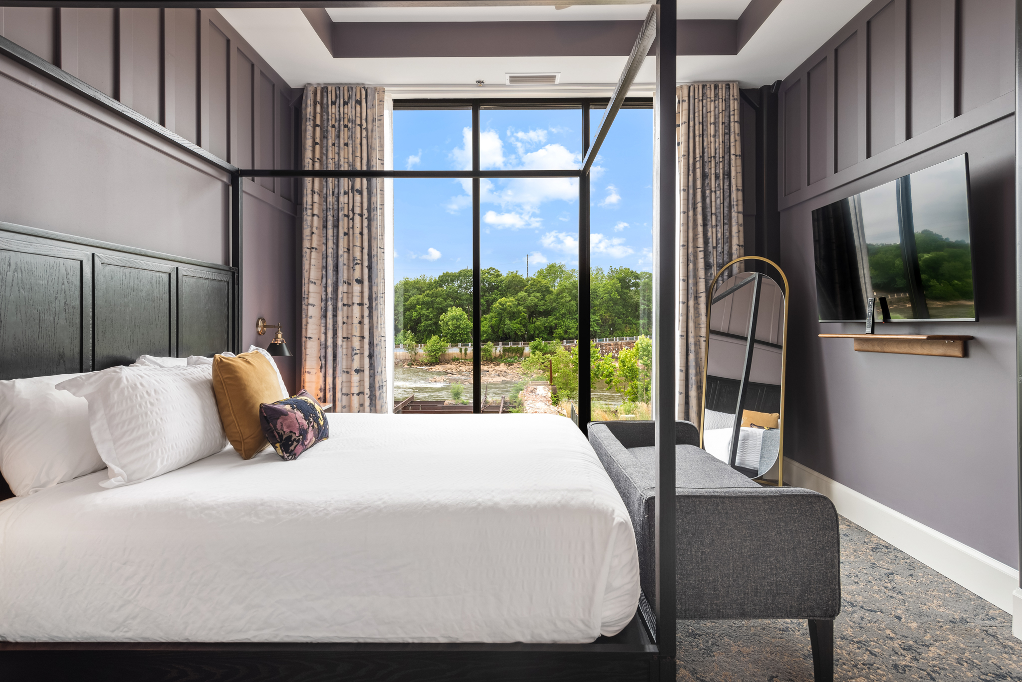 A beautifully designed hotel room featuring a four-poster bed, a cozy sitting area, large window with scenic views, modern decor, and elegant curtains.